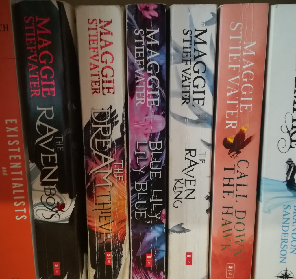 spine photograph of Maggie Stiefvater's UK editions of 'The Raven Cycle' series plus 'Call Down the Hawk. Partial showing of Iris Murdoch's 'Existentialists and Mystics' and Brandon Sanderson's 'The Final Empire', also in paperback.
