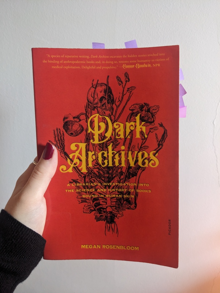 hand holding paperback of 'Dark Archives: A Librarian's Investigation into the Science and History of Books Bound in Human Skin' by Megan Rosenbloom, with paper markers